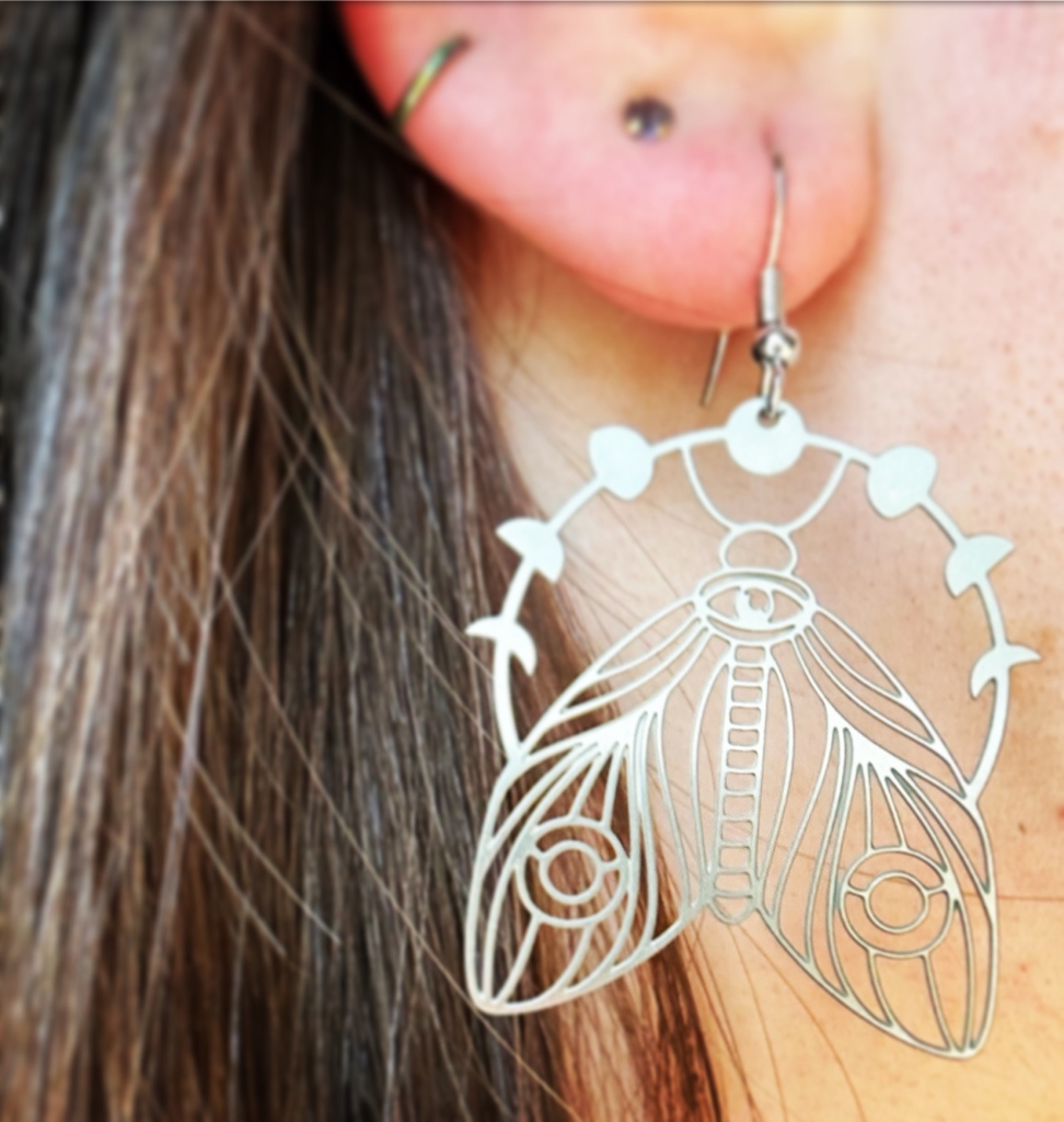 Close-up of a silver metal earring of a moth and phases of the moon, hanging from my/a brown-haired white woman's ear.